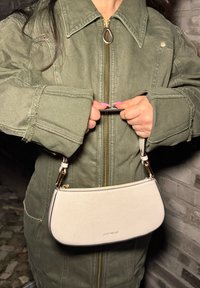 Hvitt, skinnhåndveske med buet form og gullhardware. Holdt av en person i en grønn jakke med glidelås og stor krage.