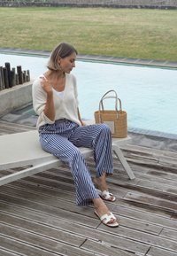 Maglione leggero beige, pantaloni a palazzo a righe blu e bianche, sandali bianchi; una borsa in rafia beige è appoggiata nei pressi di una sedia a sdraio in legno.
