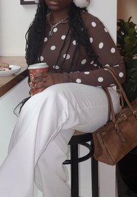 Hnědý top s puntíky a dlouhými rukávy, bílé široké kalhoty, drží kelímek s kávou, nosí stříbrný náhrdelník, sedí na černé stoličce.