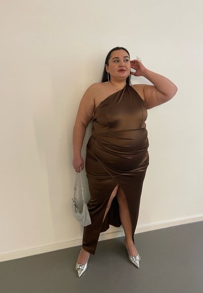 Vestido de satén marrón con un diseño de un solo hombro, textura drapeada, abertura lateral y acentos plateados. Combinado con tacones plateados de puntera afilada y bolso.