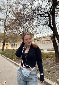 Μαύρο ριγέ φανελάκι με φερμουάρ, ανοιχτό μπλε τσάντα χιαστί, ψηλόμεσα τζιν παντελόνια, με καφέ δέντρα και κτήρια σε φόντο πάρκου.