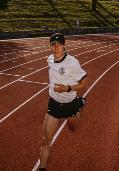 Corredor masculino con una camiseta atlética blanca y negra y pantalones cortos negros, usando una gorra negra y un reloj de pulsera, en una pista de atletismo roja.