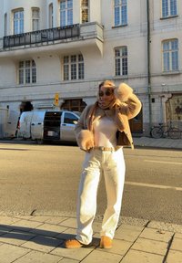 Femme portant des lunettes de soleil, une veste en fourrure beige, une écharpe, un pantalon blanc et des bottes, se tient dans une rue ensoleillée à côté d'une camionnette blanche et de vélos stationnés.