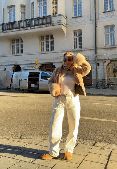 Mujer con gafas de sol, chaqueta de piel beige, bufanda, pantalones blancos y botas, de pie en una calle iluminada por el sol junto a una furgoneta blanca estacionada y bicicletas.
