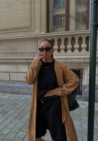 Cappotto lungo tan sopra una maglietta nera e pantaloni corti neri, accessoriato con occhiali da sole neri e orecchini a cerchio, in piedi su una strada in ciottoli.