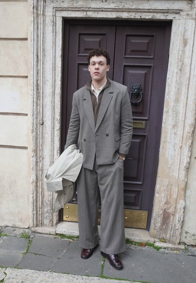 Joven con pantalones y chaqueta anchos de traje gris, de pie sosteniendo un abrigo claro frente a una puerta oscura de madera con un aldaba en forma de cabeza de león.