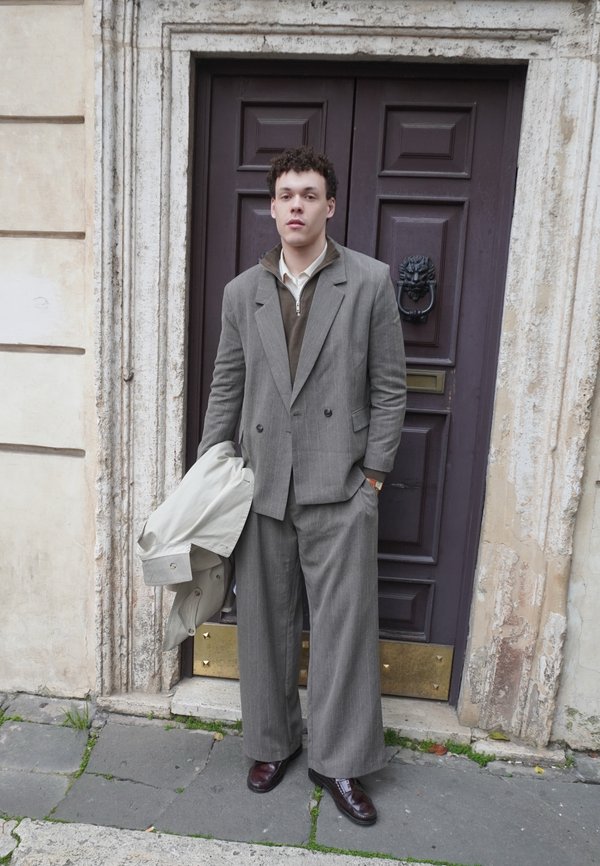 Nuori mies leveissä harmaissa pukuhousuissa ja puvuntakissa seisoo pitäen kevyttä takkia tumman puisetoven kanssa, jossa on leijonanpää-nappi.