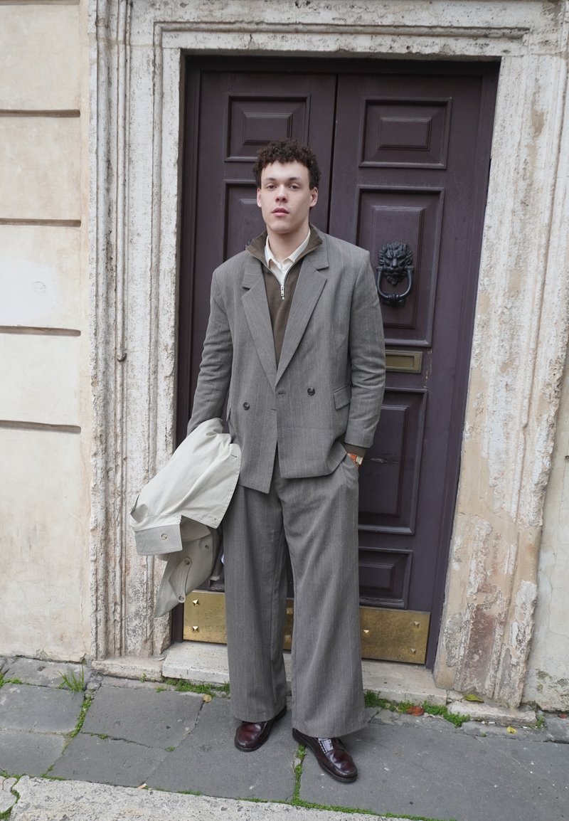 Mladić u širokim sivim hlačama i sakou stoji držeći lagani kaput ispred tamnih drvenih vrata s lavljom glavom kao kvakom.