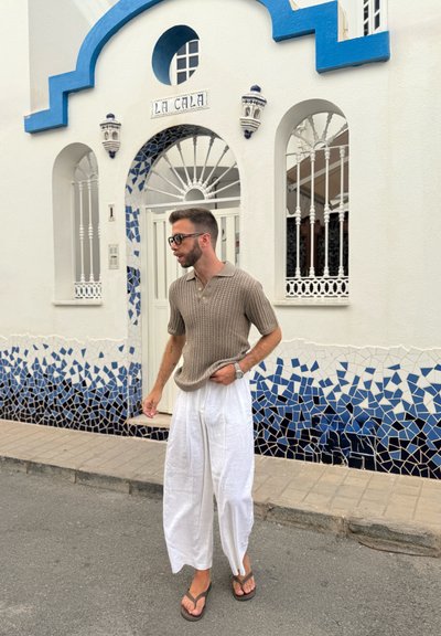 Polo de punto color caqui combinado con pantalones de lino blancos de pierna ancha y chanclas marrones, de pie contra una pared de azulejos azul y blanco.