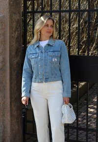 Veste en denim bleu clair courte avec poches avant et patch de logo, associée à un pantalon blanc taille haute et un sac à main tissé blanc.