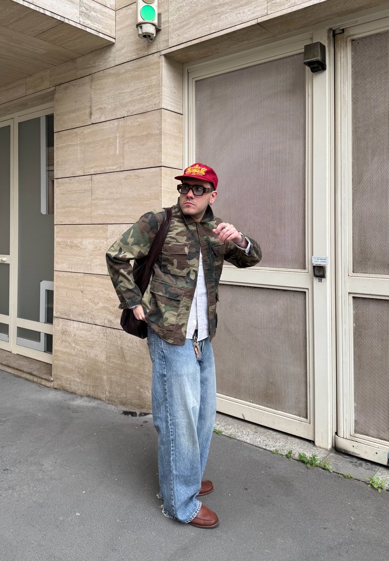 Man wearing camo jacket, red cap, sunglasses, and wide jeans stands on sidewalk near green traffic light and beige building doors.