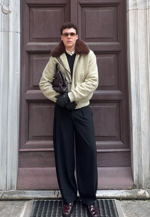 Hombre con chaqueta beige con cuello de piel marrón, guantes negros, pantalones negros de pierna ancha, gafas de sol, sosteniendo un bolso negro tejido, de pie frente a una puerta.