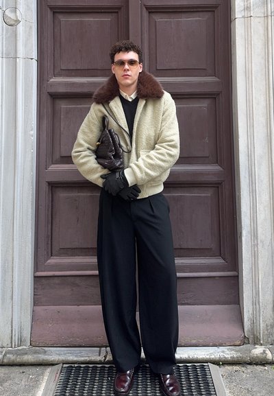 Hombre con chaqueta beige con cuello de piel marrón, guantes negros, pantalones negros de pierna ancha, gafas de sol, sosteniendo un bolso negro tejido, de pie frente a una puerta.