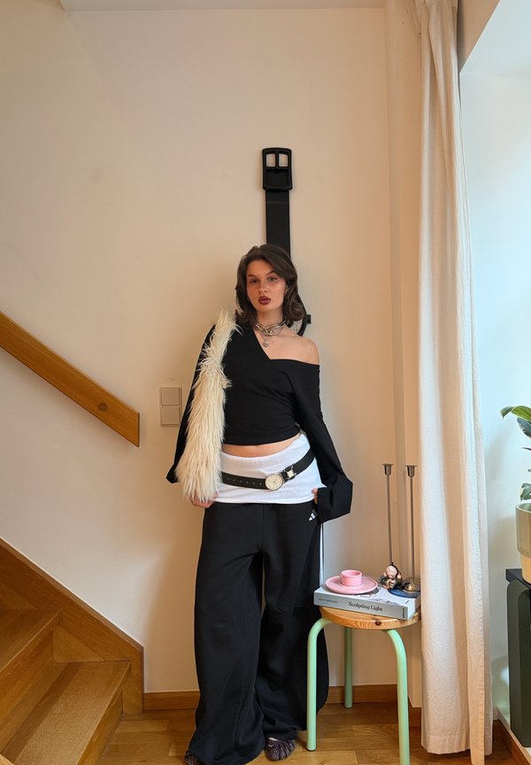 Giovane con pantaloni neri larghi, top nero scollato sulle spalle con manica bianca pelosa, in piedi all'interno vicino a una scala e un piccolo tavolo con libri.