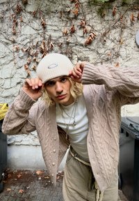 Cardigan beige a maglia con grossi bottoni sopra una t-shirt bianca, accessorato con un berretto a coste di colore chiaro. Presenta collane in strati.