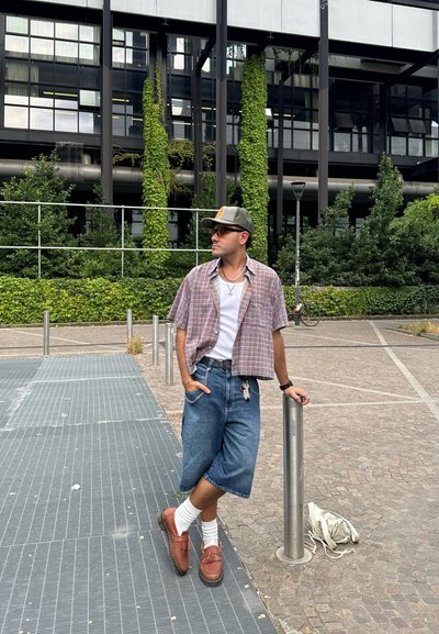 Camisa de cuadros de manga corta sobre una camiseta blanca, pantalones cortos de mezclilla, zapatos marrones y calcetines blancos. Los accesorios incluyen un reloj y una cadena.