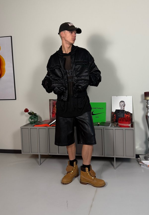 Casaco bomber de cetim preto sobre uma camisa de malha preta, combinado com calções de couro pretos e botas de trabalho castanhas. Inclui boné de beisebol e acessórios.