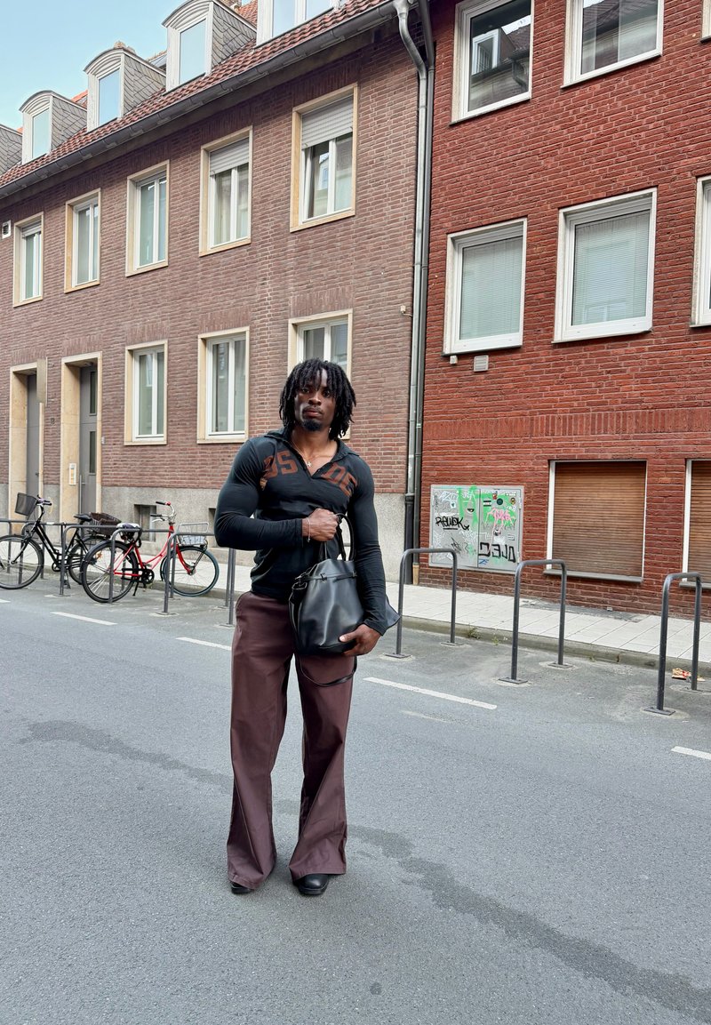 Uomo con dreadlocks in piedi per strada, tiene una borsa nera, indossa una camicia nera a maniche lunghe e pantaloni larghi marroni, con edifici in mattoni sullo sfondo.