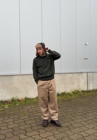 Zöld pulóver, bézs nadrág és barna szőrmebéléses kalap, macskakövön állva, háttérben egy texturált szürke fal.