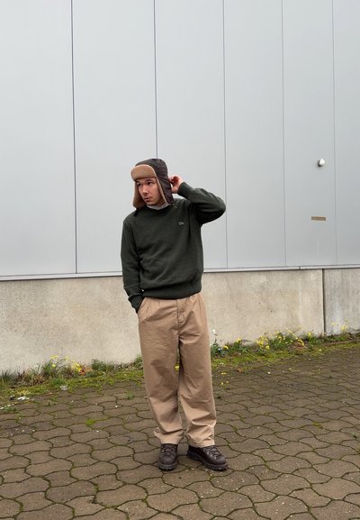 Suéter verde, pantalones beige y sombrero marrón forrado de piel, de pie sobre adoquines, con una pared gris texturizada en el fondo.