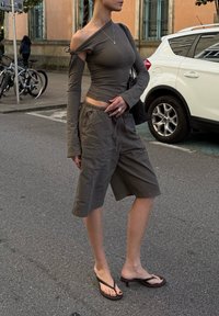 Camiseta de manga larga en gris oscuro con diseño de hombros al descubierto, combinada con pantalones cortos cargo grises sueltos y sandalias de tacón flip-flop negras. Tejido texturizado y ajuste casual.