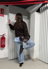 Femme en pull noir duveteux, jean bleu clair et chaussures noires, debout sur une jambe près d'un extincteur rouge dans un espace industriel blanc.