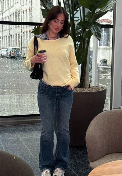 Mujer con suéter amarillo y jeans sosteniendo una taza de café y un bolso negro, de pie en el interior cerca de grandes ventanas con una planta en maceta.