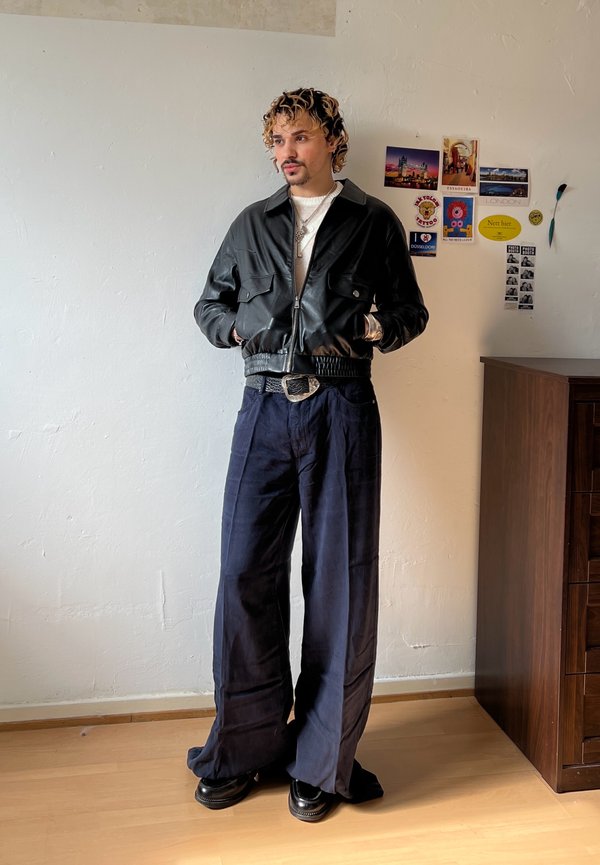 Joven con cabello rizado lleva una chaqueta de cuero negra, pantalones anchos azul marino y zapatos negros, de pie en una habitación con postales en la pared.