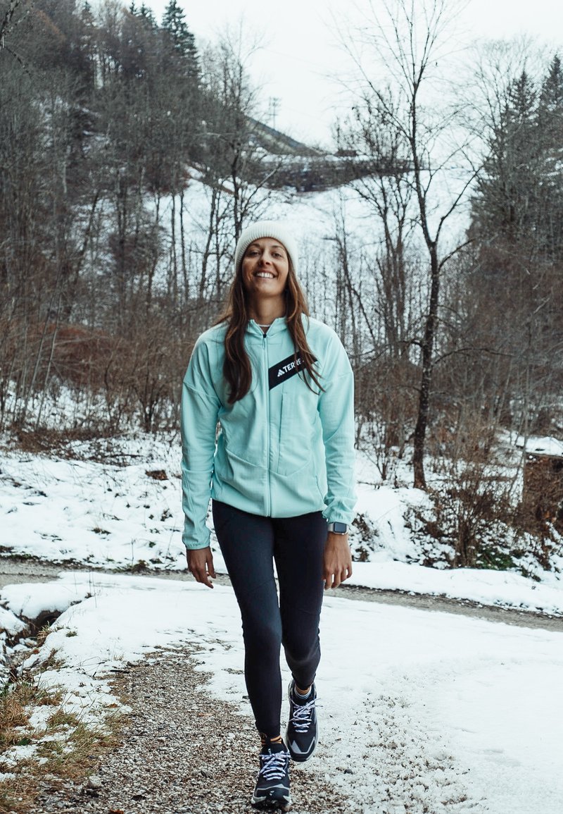 Vrouw in een lichtblauwe jas en witte muts wandelt op een besneeuwd pad met bomen en heuvels op de achtergrond, ze glimlacht en draagt zwarte leggings.