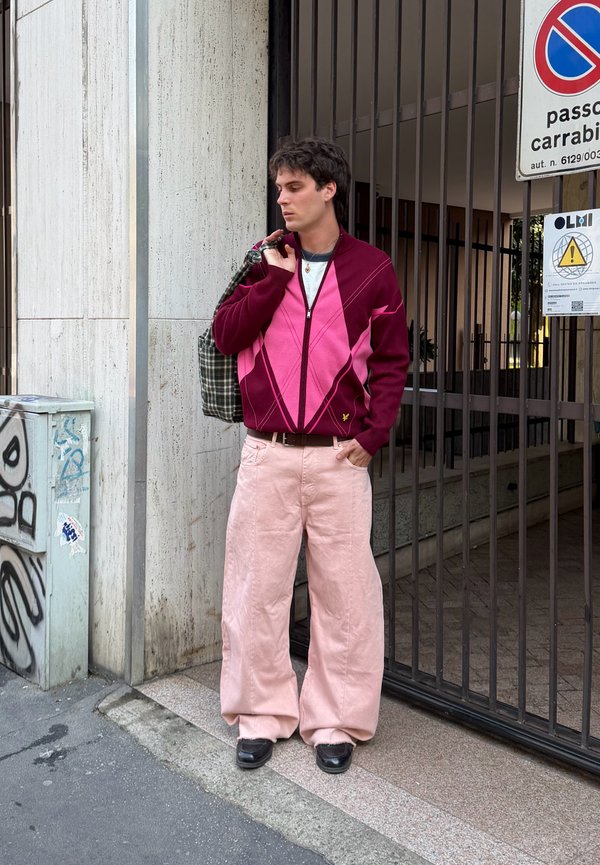 Jeune homme en pull rose et bordeaux, pantalon large rose clair, chaussures noires, portant un sac à carreaux, debout près d'une grille en métal et des panneaux de rue.