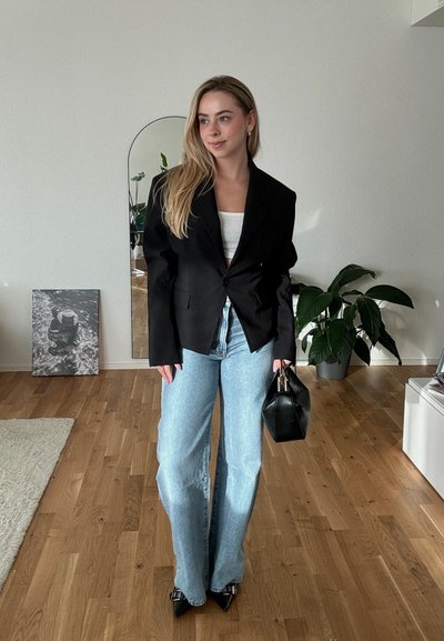 Blazer negro a rayas con dos bolsillos frontales, combinado con jeans de pierna ancha de color azul claro. Sostiene un pequeño bolso negro, de pie sobre un suelo de madera.