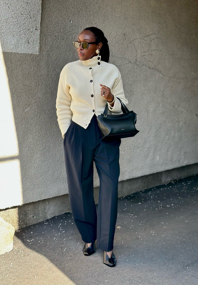 Kvinne iført kremfarget knappet cardigan, marineblå bukser, svarte hæler, solbriller, holder en svart veske, står mot en teksturert grå vegg.