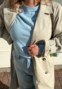 Světle béžový trenčkot přes světle modrou košili, doplněný světle modrými džíny. Výrazné detaily zahrnují zlatý prsten a vzorovanou tašku.