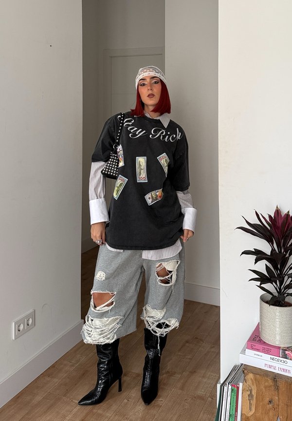Camisa negra oversized con texto "Stay Rich", pantalones cortos desgastados grises, botas altas negras, camisa blanca de cuello por debajo. Pequeño bolso negro con tachuelas.