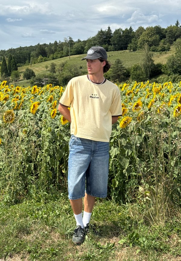 Jonge man met pet, geel t-shirt en spijkershorts staat met handen achter zich op een zonnebloemveld onder een bewolkte hemel.