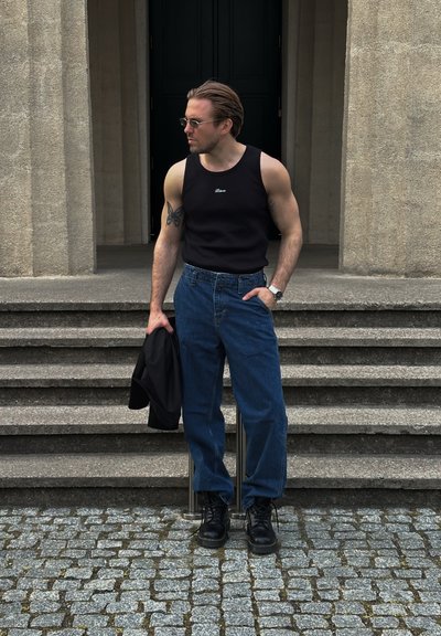 Camiseta sin mangas negra con logo, jeans ajustados de tiro alto azules, botas negras y gafas de sol. De pie en escaleras de piedra. Sosteniendo una chaqueta negra.