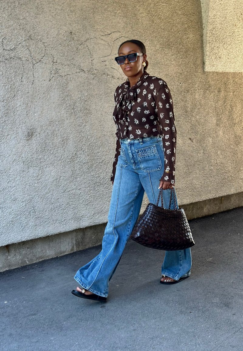 Bluse mit braunem Blumenmuster und gebundenem Kragen, hochgeschnittene blaue Schlagjeans und eine große schwarze geflochtene Handtasche. Glatte Flip-Flops.