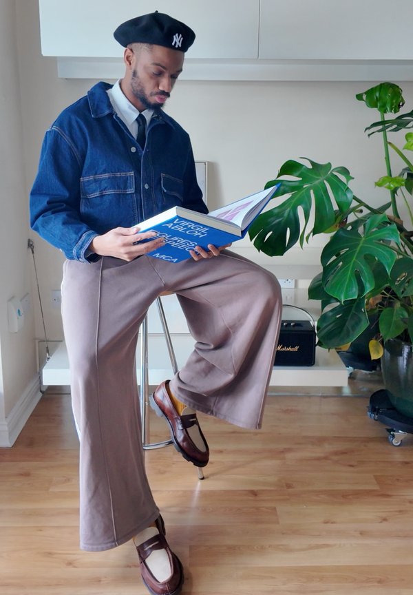 Mand i denimjakke og beret sidder på en skammel og læser en stor blå bog, ved siden af en stor potteplante indendørs.