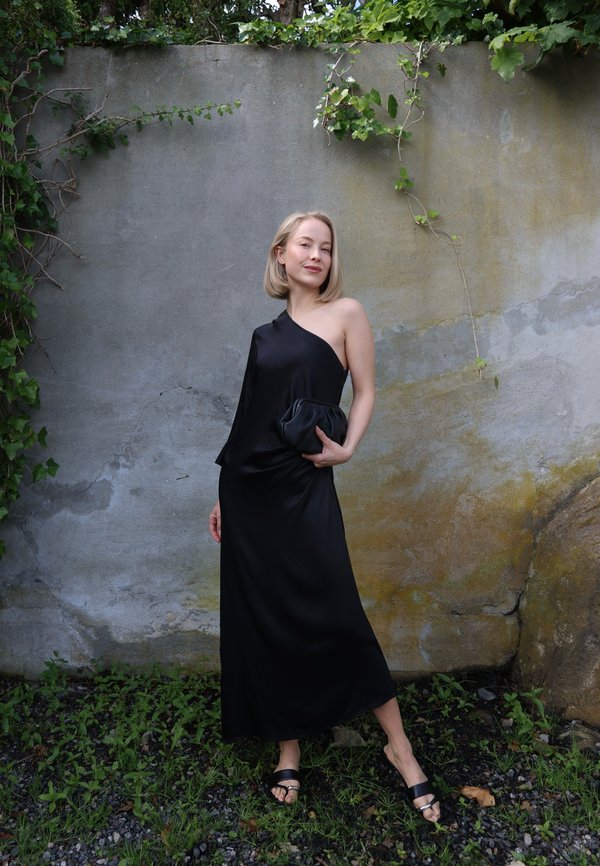 Frau mit kurzen blonden Haaren, die ein ein- schulteriges schwarzes Kleid und Sandalen trägt, steht vor einer verwitterten Wand mit grünen Ranken.