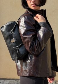 Crna kožna torba na leđa s elegantnim dizajnom, srebrnim dijelovima i kopčama, nošena preko sjajne smeđe jakne s definiranim ramenima.