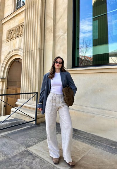 Blazer gris de gran tamaño, camiseta sin mangas blanca, pantalones de lino beige de pierna ancha y bolso marrón texturizado. El fondo presenta arquitectura de piedra y grandes ventanales.