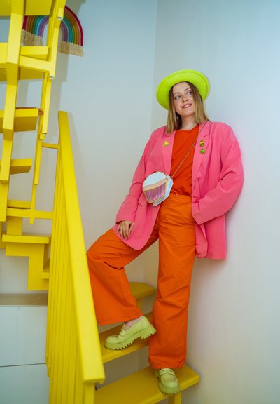 Mujer joven con pantalones naranja brillante, camiseta roja, blazer rosa y sombrero verde de pie en una escalera amarilla con un bolso en forma de cupcake.