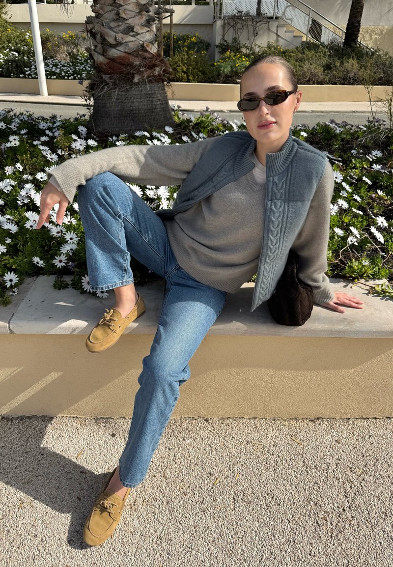 Woman wearing sunglasses, a grey sweater vest, and jeans, sitting on a ledge beside white flowers and a palm tree in daylight.