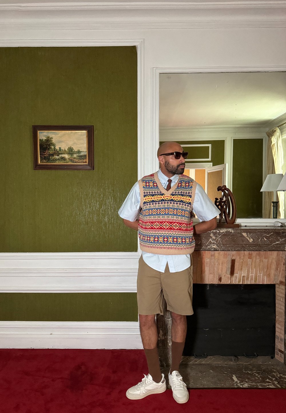Πλεκτό γιλέκο με πολύχρωμα σχέδια πάνω από ανοιχτό μπλε πουκάμισο, συνδυασμένο με μπεζ σορτς και λευκά αθλητικά παπούτσια, σε φόντο πράσινου τοίχου.