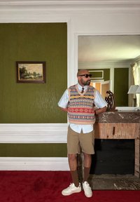 Πλεκτό γιλέκο με πολύχρωμα σχέδια πάνω από ανοιχτό μπλε πουκάμισο, συνδυασμένο με μπεζ σορτς και λευκά αθλητικά παπούτσια, σε φόντο πράσινου τοίχου.