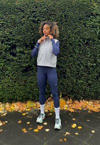 Pelēks veste virs zila garām piedurknēm tops, apvienots ar ziliem legingiem un gaiši zaļām skriešanas kurpēm. Stāv uz ceļa ar rudens lapām.