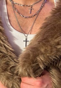 Faux-Fur-Braunjacke mit strukturierter Oberfläche, geschichteten Silberketten und einem zentralen Kreuzanhänger über einem weißen, gerippten Tanktop.