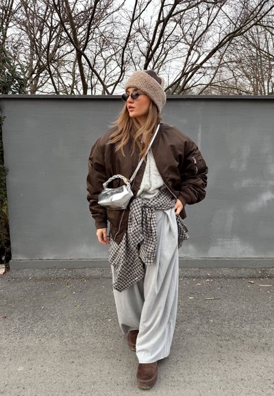 Mujer con chaqueta bomber marrón, gorro de punto beige, gafas de sol redondas, pantalones anchos grises, con una camisa de cuadros atada a la cintura y un pequeño bolso plateado.