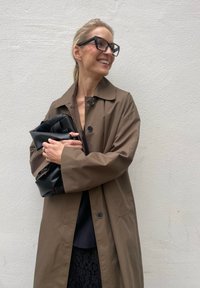 Brown oversized trench coat with large buttons, worn over a black top and skirt. Holding a pleated black bag against a white wall backdrop.