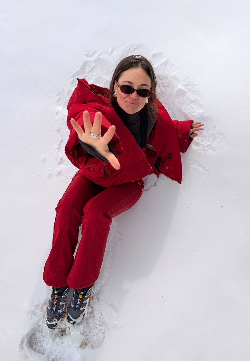 Femme en tenue d'hiver rouge et lunettes de soleil assise dans la neige, tendant la main vers la caméra avec une expression joueuse.